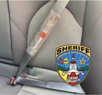 Seatbelt with a gray cover labeled “Autism” and a red autism awareness puzzle piece, shown beside the Leon County Sheriff badge with the phrase “Embrace Differences.”
